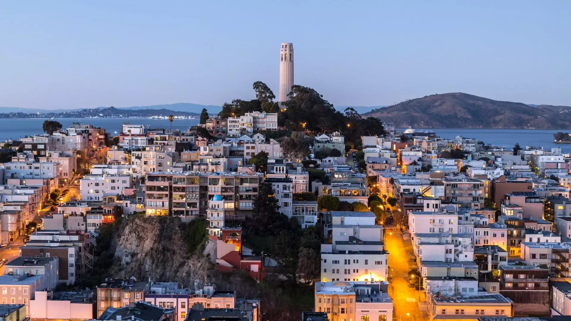 Coit Tower de San Francisco al anochecer, con calles iluminadas frente a ella y la Bahía de San Francisco detrás.