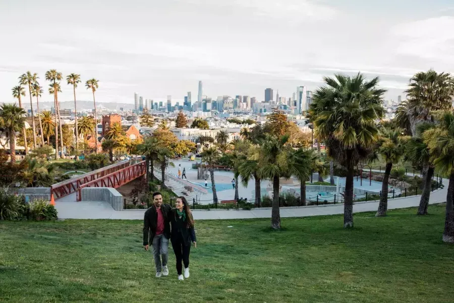 Una pareja camina hacia la cámara con Dolores Park y el Skyline de San Francisco detrás de ellos.